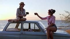tea party of Girls with cups into Hands at roof and bonnet vehicle on seafront in sundown,