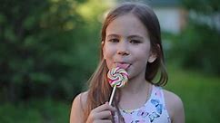Close-up front view of pretty brunette Caucasian girl with brown eyes looking at camera licking multicolored lollipop in slow motion. Happy satisfied child posing in summer park enjoying tasty dessert