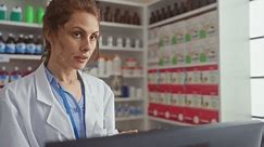 Une pharmacienne concentrée travaille sur un ordinateur dans une pharmacie avec des rayons de médicaments derrière elle.