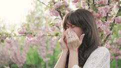 Sick brunette woman having runny nose and suffering from seasonal allergy near blooming sakura tree