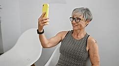 A smiling mature woman with grey hair takes a selfie indoors with a yellow phone, exuding confidence and joy.