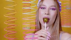 Sexy girl smiles, licks round caramel on stick in room with yellow wall, slinkies hanging from ceiling. Frisky blonde with lollipop. Playful woman holds, puts candy in mouth. Vibrant saturated colors