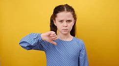 Portrait of displeased little girl child 8-9 years old wearing blue dress show thumb down dislike gesture, posing isolated over yellow color background wall in studio. Childhood lifestyle concept