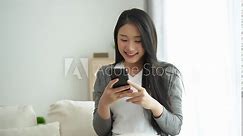 Asian woman hold mobile phone text or message sitting on coach sofa in the living room. Woman smiling use smartphone to browse web, read e-book, trade stocks at home.
