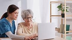 Caregiver helping retired senior woman use computer. Friendly young nurse caretaker girl showing older lady how to do web search, send emails, watch videos and open digital telemedicine app on laptop