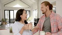 A diverse couple shares a cozy moment in a modern kitchen at home. The Caucasian male embraces his Asian girlfriend, both smiling, as she holds a mug, unaltered, slow motion.