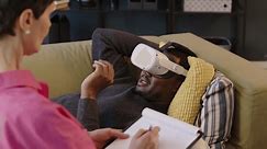 Black male patient wearing vr headset lying on sofa at modern psychotherapist office and explaining his feelings to female specialist sitting nearby with clipboard for note taking
