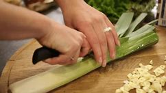 Knife skills, Precision chopping, Slicing leeks. Hands expertly wielding knife, chopping leek on wooden cutting board, with sliced garlic and fresh vegetables scattered around.