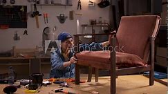 Woman in a workshop inspecting an old wooden chair before restoration. Tools and equipment are visible on the workbench and walls, creating a scene of furniture renovation preparation.