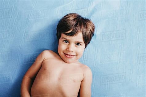 From above of cheerful shirtless little boy looking at camera while lying on blue hospital bed 