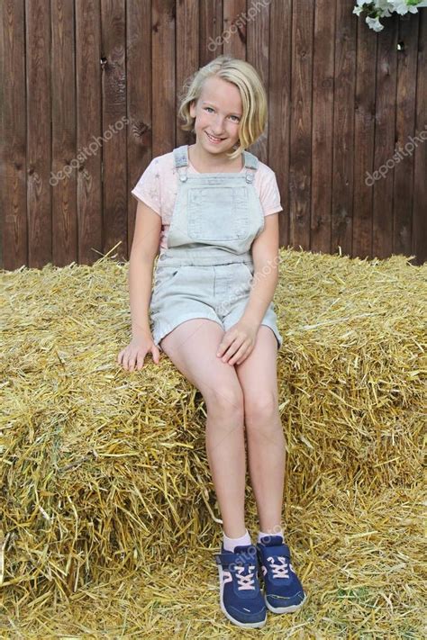 The specified thread does not exist. Beautiful happy preteen girl dressed in short bib overalls sitting on a hay in the village ...