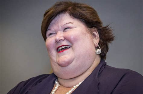 Maggie looking sideways, short brown hair, pink scarf, black vest, quote in white letters above her head: Maggie De Block is Belgium's 20 stone Minister of Public ...