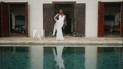 Non-binary black person model poses in white dress at luxury villa entrance by swimming pool. Gay black man raises hands up, touches transparent veil and reflects in blue water. Destination wedding.