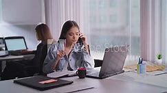 Asian India teen staff girl sitting desk chair use laptop hold mobile phone talk friend client help online indoor office Indian busy young adult gen z woman lady digital call device do job work task