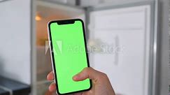 Woman's hand scrolls touch screen mobile phone with green chromakey against background of an open empty refrigerator, making purchases, looking at prices in app.