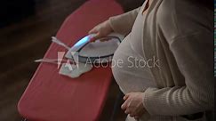 An unrecognizable pregnant woman steams and irons white knitted booties with a steam iron on a red ironing board, standing indoors