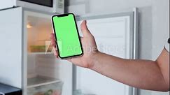 Mobile phone with green screen display in hand, by scrolling finger to touch screen, against the background of an open empty refrigerator, in kitchen.
