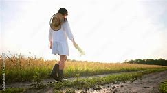 Brunette woman in airy white dress, sun hat and rubber boots with bouquet of field flowers walking along country road between agricultural fields in village, back view. Carefree, romantic, rural life.