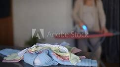 In the foreground there is a stack of baby clothes on the table. In the background, out of focus, an unrecognizable pregnant woman irons clothes on an ironing board with a steam iron while standing in