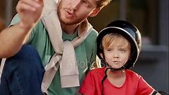 A dad with curly hair in a Green T-shirt and blue jeans sits with his little blond son in a black safety helmet and a red T-shirt on the curb in a park near a building and looks at something around