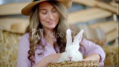 Woman petting a white bunny in the barn near square hay bales, in daylight