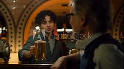 Young bartender pours craft beer into glass and gives it to mature man. Male friends clink glasses and drink beer sitting at the bar counter in stylish pub at night. Concept of lifestyle and leisure.