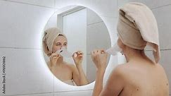 Winsome beautiful Caucasian woman wrapped in towel on her head looking at mirror at her reflection removing cloth mask doing home cosmetology procedures standing in bathroom