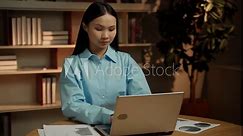 A focused young Asian woman is diligently working at her desk in a contemporary office setting, using her laptop while reviewing documents and charts.